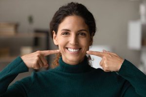 Smiling woman in a green turtleneck pointing at her teeth, showcasing a healthy smile, relevant to dental crown restoration and oral health.